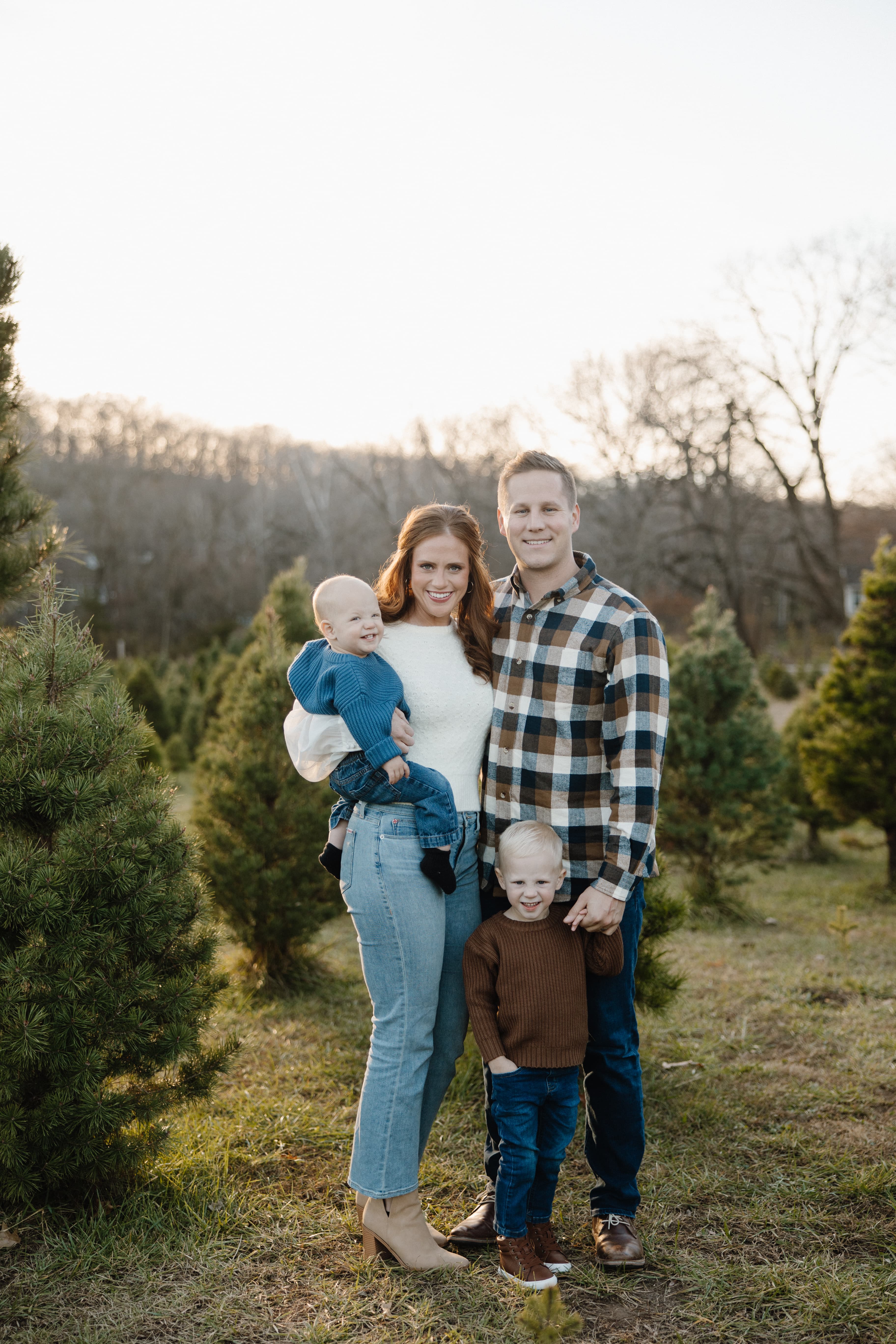 Smiling family of four standing together in a field of evergreen Christmas trees.